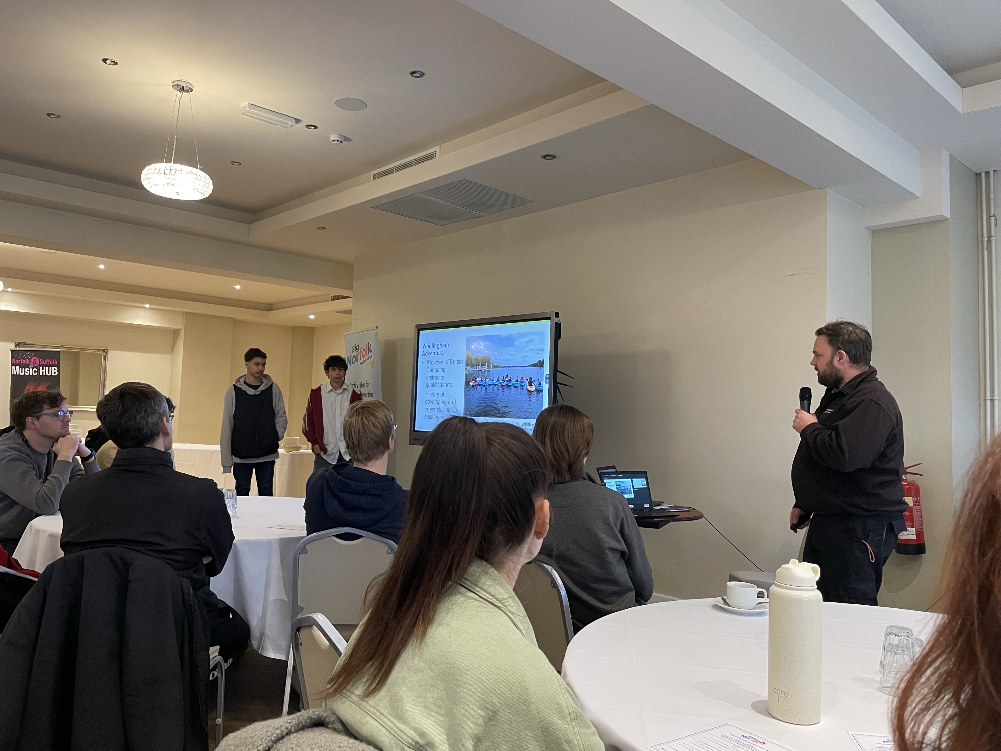 Person speaking at function with others sitting around looking at slide show on screen