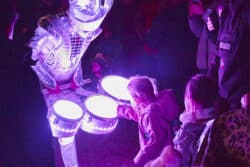 children looking intently at fluorescent drums carried by actor in silver suit